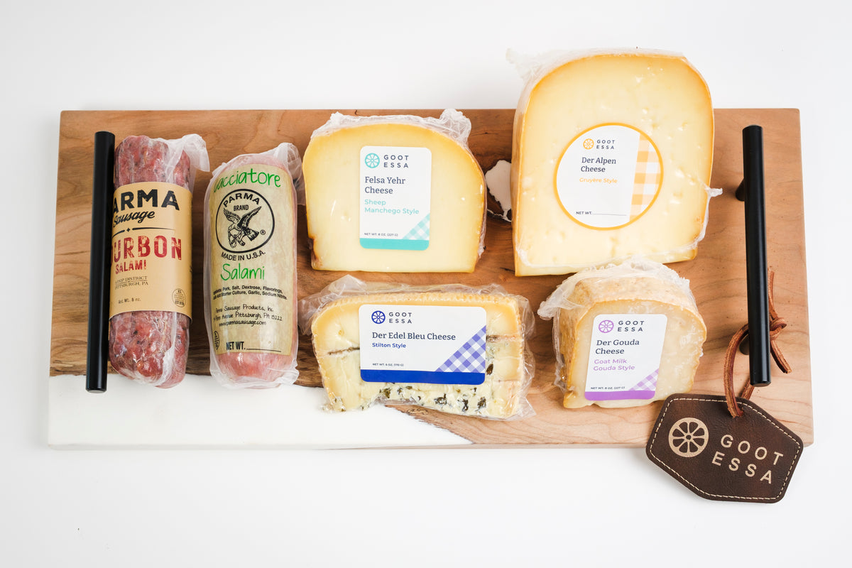 A curated cheese and cured meat charcuterie board arranged on a wooden board with a white marble runner. The assortment includes Gootessa cheese wheels and blocks, a salami roll, a salami stick, and a salami slice, with two black picks on either side and a Gootessa brand tag hanging from the right side.