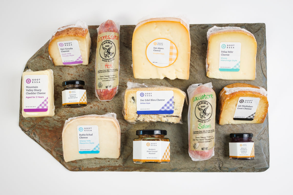 An assortment of artisanal cheeses and spreads arranged on a rustic dark stone serving board against a clean white background. The products include various cheeses with colorful labels and small jars of spreads, branded with the logo 'The Cheese Co.
