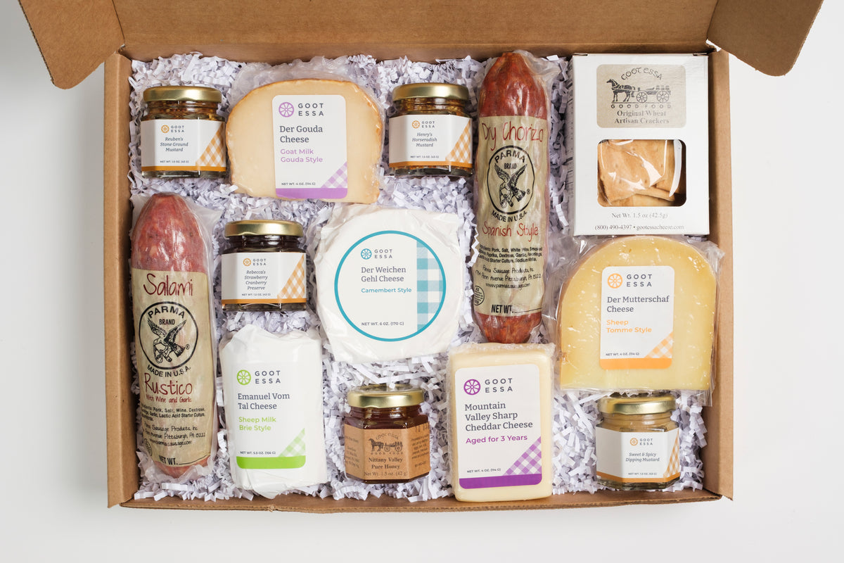 An open cardboard gift box filled with assorted cheeses, spreads, and packaged food items on white shredded paper. Visible brands include Good Grass and Saloon.